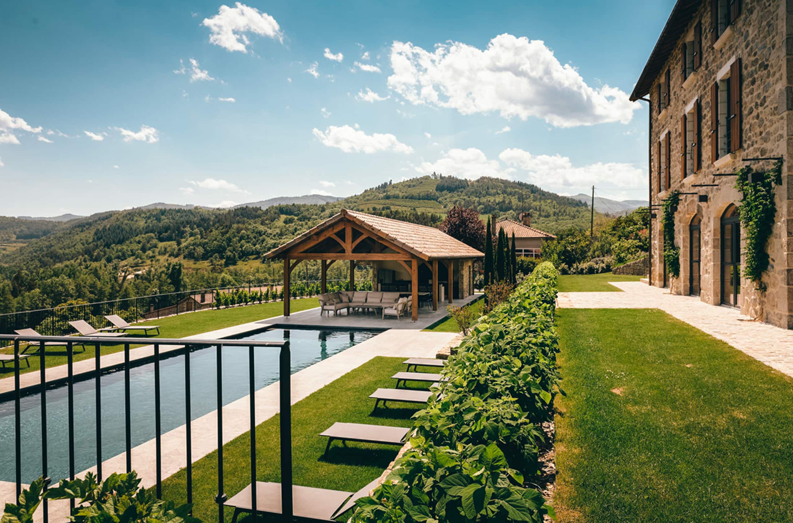 A large stone building with an infinity pool set in a landscaped garden, overlooking a valley of green hills beneath a clear blue sky dotted with clouds.