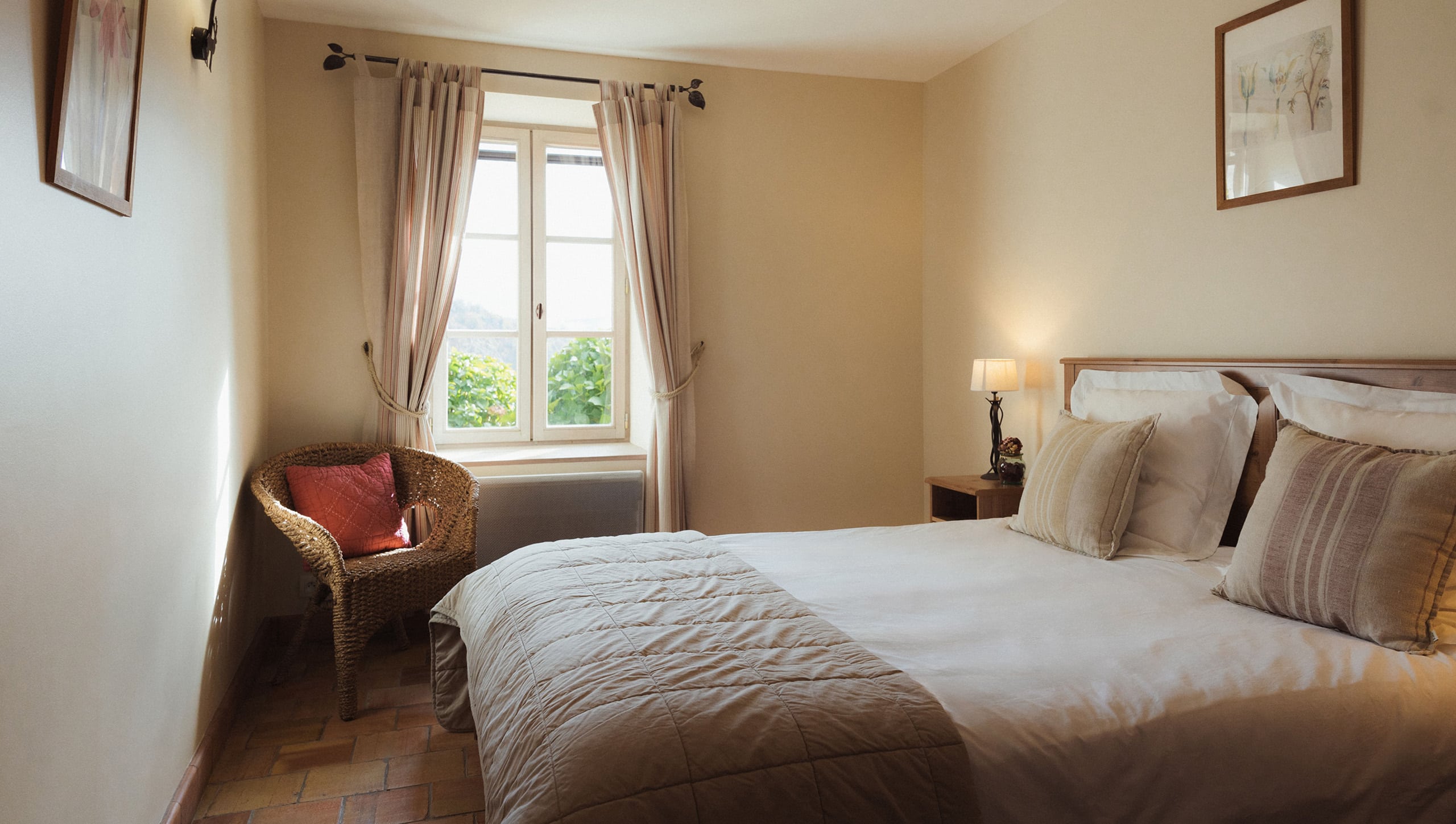 Chambre d'hôtel lumineuse : lit fait avec coussins, chaise en osier près d'une fenêtre ouverte sur la verdure. Murs clairs et sol en terre cuite.