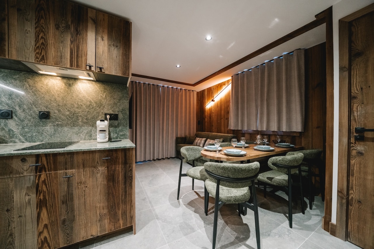 Intérieur d'appartement moderne: cuisine en bois rustique avec comptoir marbré, adjacente à une table à manger dressée pour six personnes et des chaises vertes. Murs lambrissés, rideaux beiges.