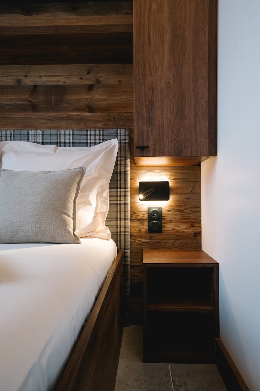 Un lit double est préparé avec une tête de lit écossaise et des coussins blancs. Une lampe murale illumine une table de nuit en bois, sur un mur lambrissé de bois foncé.