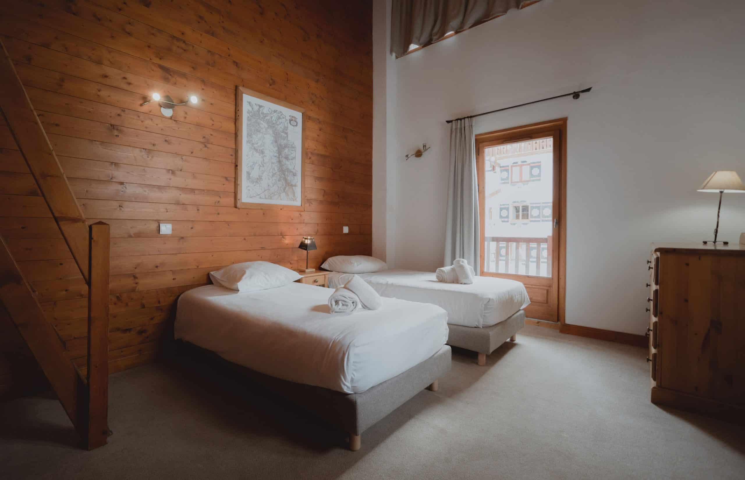 Deux lits simples sont préparés avec des serviettes roulées, dans une chambre aux murs en bois, sous une carte murale et face à un balcon donnant sur des bâtiments.