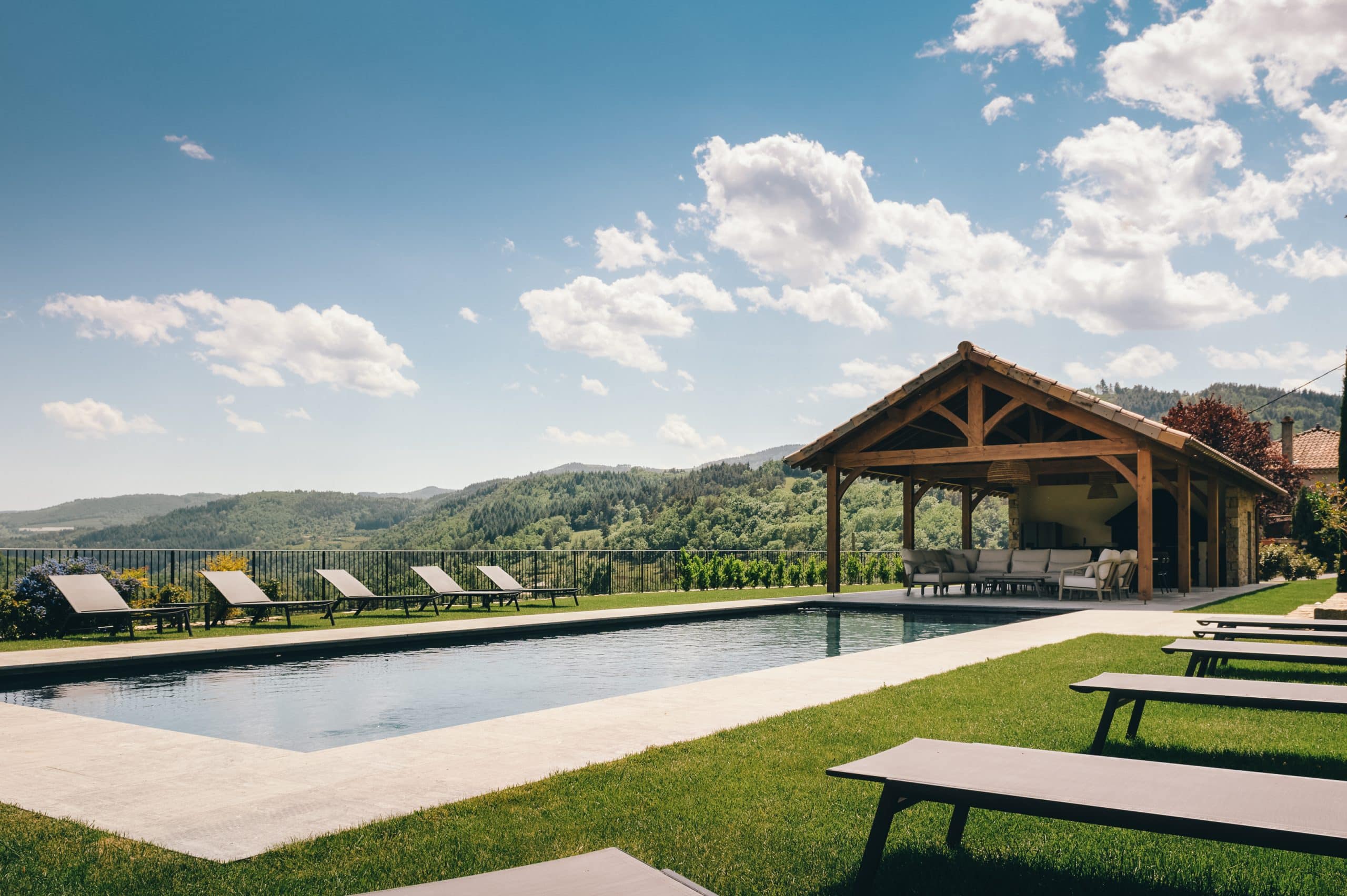 Piscine moderne et salon extérieur sous charpente en bois, aménagés pour la détente, surplombent un paysage montagneux verdoyant sous un ciel bleu nuageux.