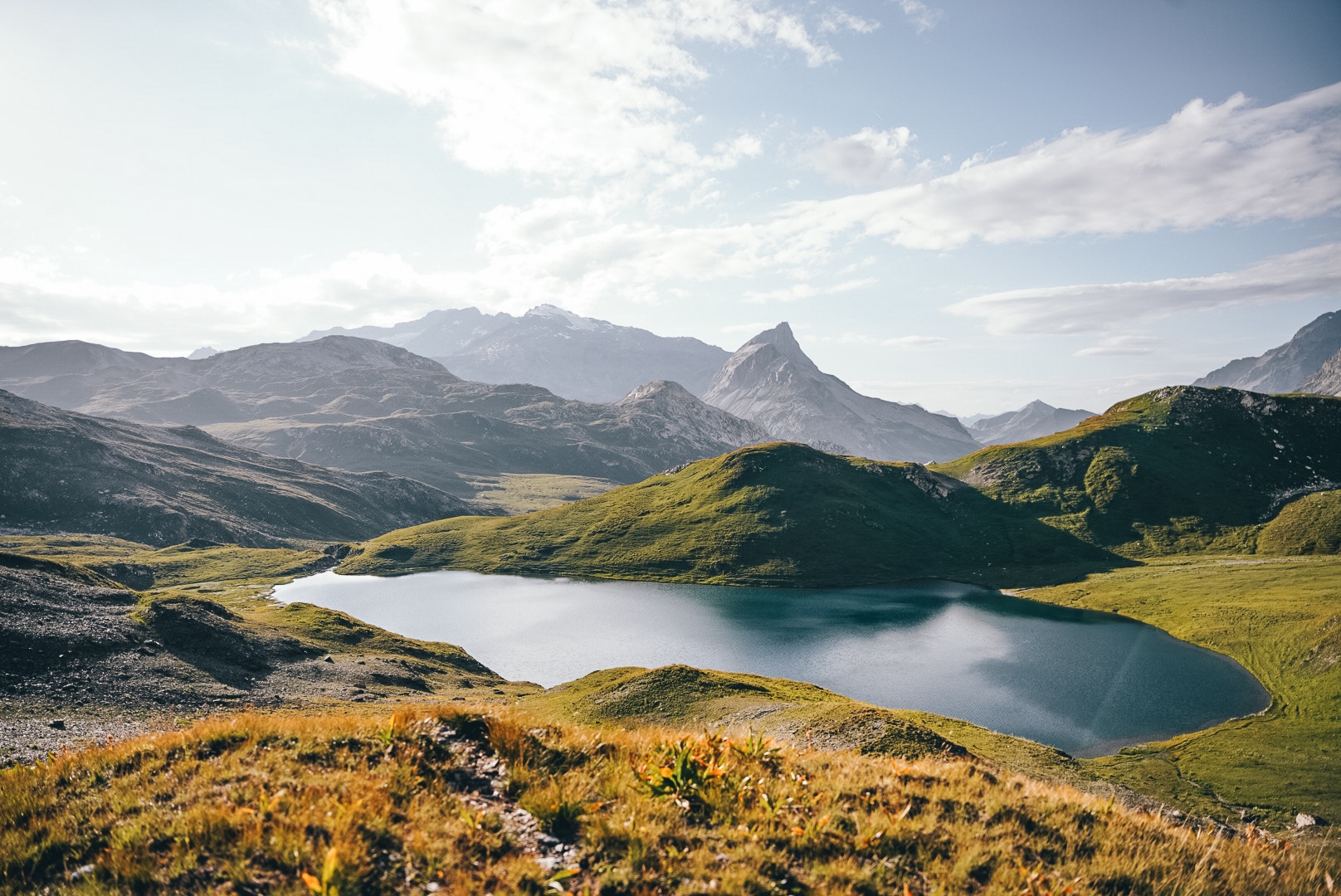 Un lac de montagne aux eaux calmes est niché entre des collines verdoyantes et des sommets rocheux escarpés, sous un ciel partiellement nuageux, avec de l'herbe sèche au premier plan.