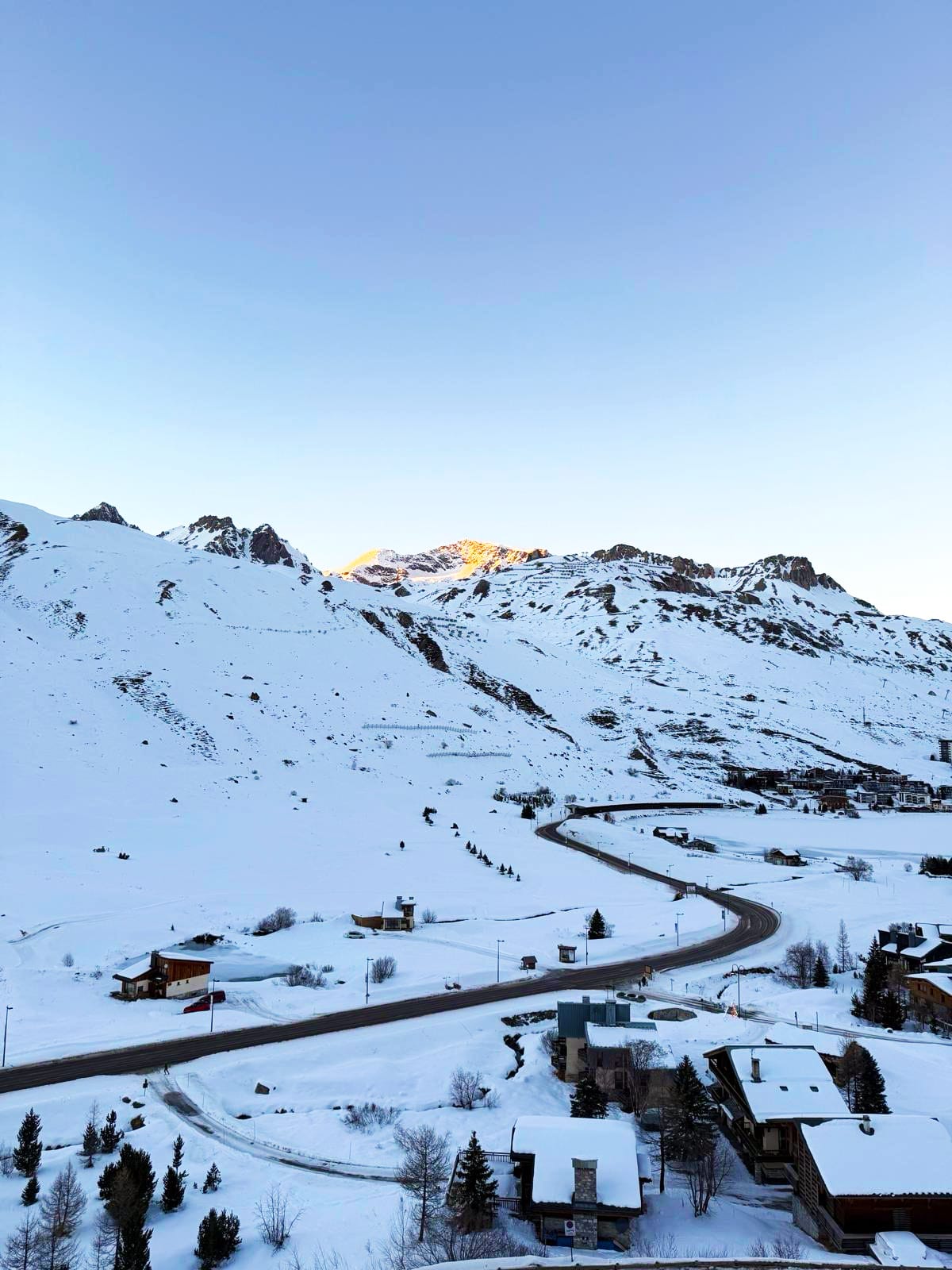 Une route sinueuse mène à travers un village de montagne enneigé. Des chalets aux toits blancs parsèment le paysage, avec des sommets lointains dorés par le soleil sous un ciel clair.
