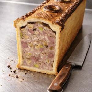 Pâté en croûte doré, coupé en deux, révèle une farce de viande rose, pistaches et jambon. Il repose sur une surface métallique près d'un couteau et de grains de poivre.