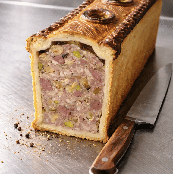 Pâté en croûte doré, coupé en deux, révèle une farce de viande rose, pistaches et jambon. Il repose sur une surface métallique près d'un couteau et de grains de poivre.